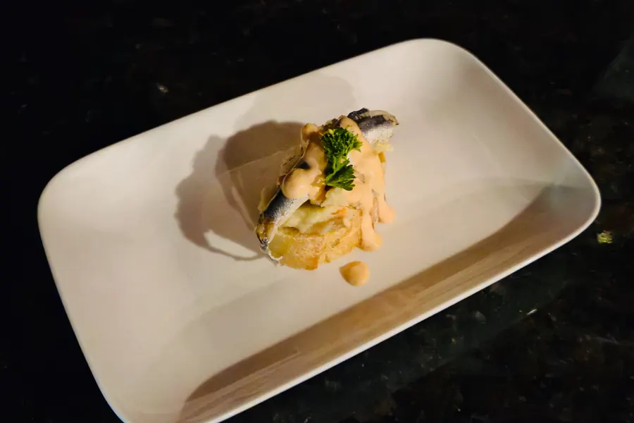 A sardine pincho at La Vid: a sardine fillet on bread with sauce and parsley, on a white plate