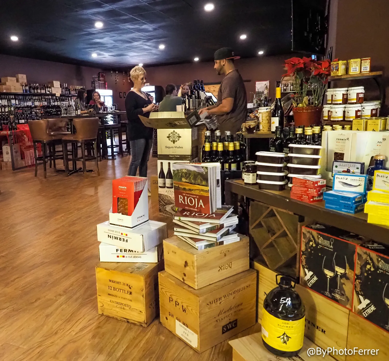 Inside La Vid: guests at the register, leather bar stools and a high table, wine walls along both sides, and crates of port, Fermín ibérico, Rioja books, and tinned fish stacked in the foreground