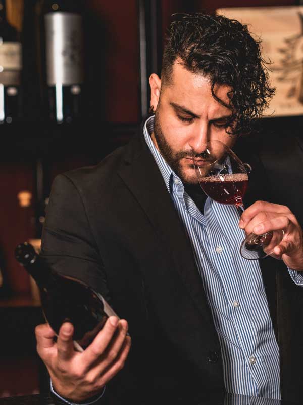 Victor Manuel nosing a glass of red wine while reading the label of the bottle in his other hand
